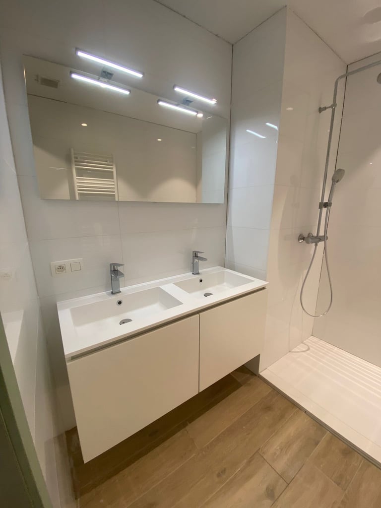 Moderne badkamer met dubbele witte spoelbak, verlichte spiegel, houten vloer en inloopdouche met glazen cabine